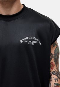 Zwarte korte mouwen, mesh T-shirt met een ronde halslijn met daarop witte geborduurde tekst: "GOOD FOR NOTHING BRITISH ATELIER EST 2011."