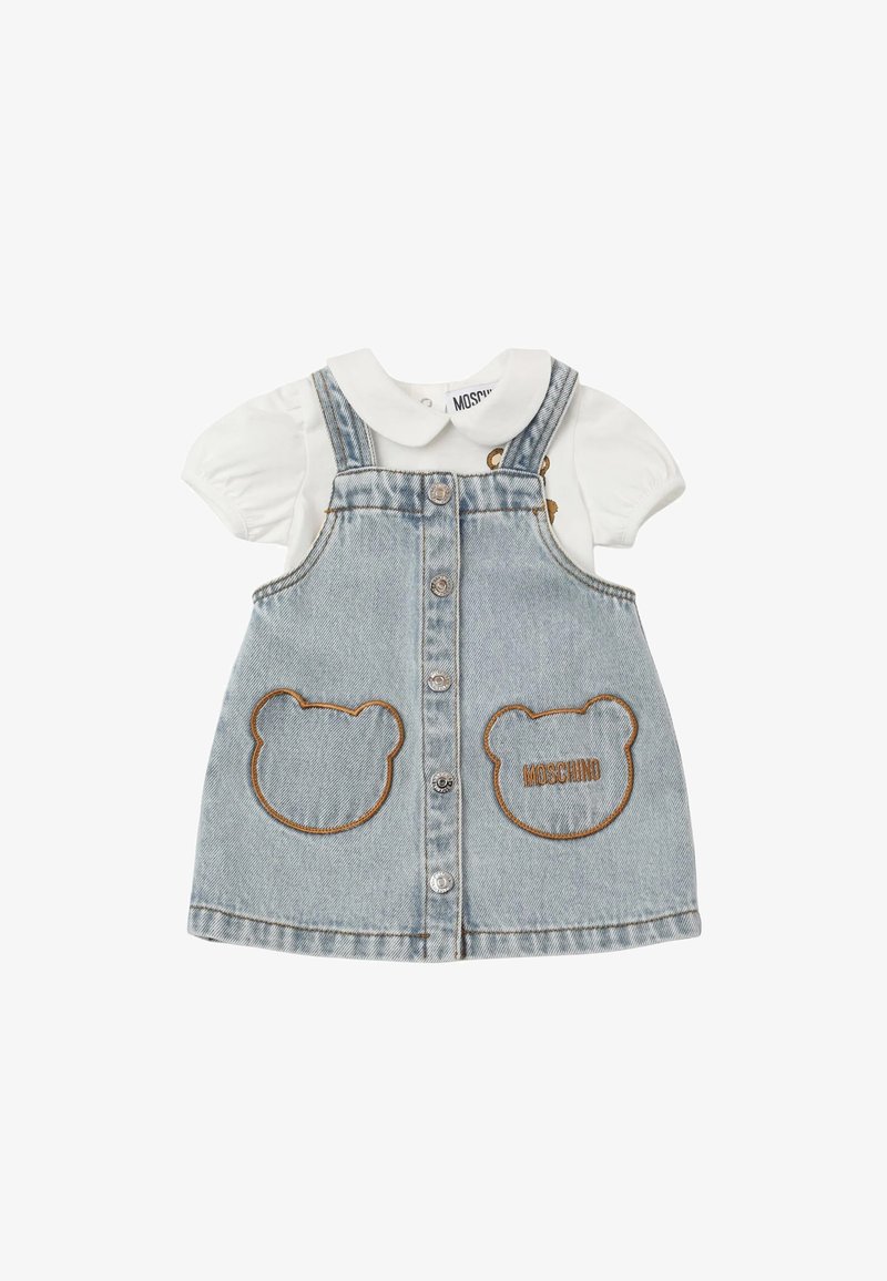 Abito da salopette in denim con un lavaggio blu chiaro, caratterizzato da tasche a forma di orso e ricamo "Moschino"; camicia bianca con maniche a sbuffo e colletto sotto.