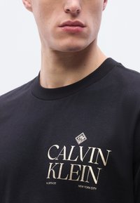 Μαύρο βαμβακ ερό μπλουζάκι με χρυσό λογότυπο που γράφει «CALVIN KLEIN» και «W. 39TH ST. NEW YORK CITY». Στρογγυλή λαιμόκοψη, κοντά μανίκια.