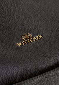 Superficie in pelle nera con texture che presenta un logo "WITTCHEN" in tono dorato con un emblema sopra di esso, che mette in mostra un design embossato.