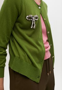 Grön cardigan i mjukt material, med en smyckad brosch på vänster sida, bärs över en rosa topp och matchas med bruna byxor.