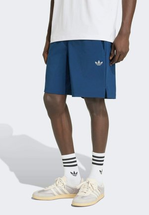 Mężczyzna w niebieskich szortach Adidas z logo, białych skarpetach Adidas z czarnymi paskami i beżowych butach sportowych Adidas na białym tle.