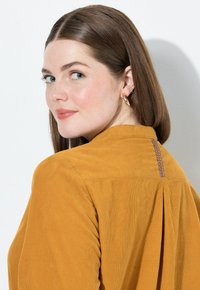 Senfgelbe Cordshirt mit gefälteter Rückenpartie, einem einfachen Kragen und einer kontrastierenden Naht entlang des Halsausschnitts.