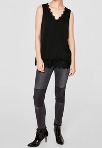 Černý bezrámový top s krajkovým lemem a výstřihem do V, doplněný tmavě šedými skinny džínami s kontrastními látkovými panely a lesklými kotníkovými botami.