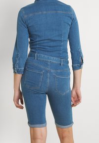 Person som bär en åtsittande medelblå denimjacka och shorts med rullade fållar, står vänd bort från kameran mot en enfärgad bakgrund.
