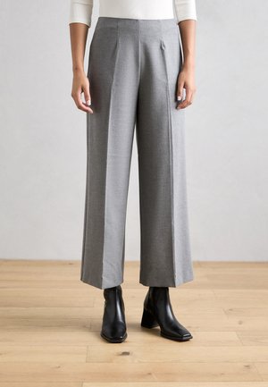 Weite Stoffhosen aus strukturiertem grauem Material mit Vorderfalten, hoher Taille und kombiniert mit schwarzen Knöchelstiefeln auf einem Holzboden.