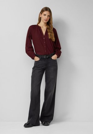 Bordeauxrote Bluse mit langen Ärmeln und Knopfleiste vorne, kombiniert mit hoch taillierten, weit geschnittenen schwarzen Jeans und schwarzen Plateauschuhen. Ein Gürtel betont die Taille.