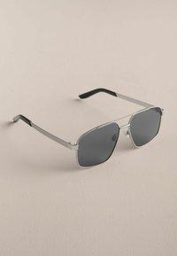 Lunettes de soleil à monture en métal argenté avec des verres rectangulaires foncés et des branches noires, posées sur une surface beige clair.