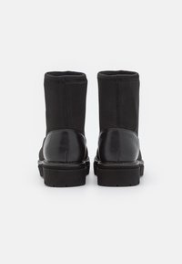 Par de botas negras de media caña con suelas de goma gruesa y secciones de tacón de cuero, mostradas desde atrás sobre un fondo blanco.