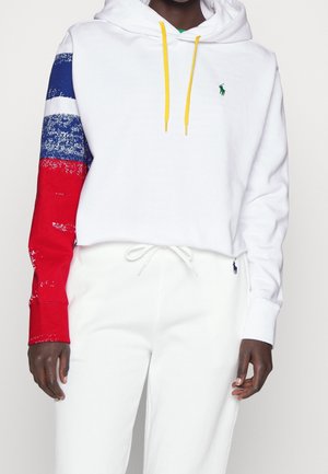 Person iført hvid croppet hættetrøje med gule snore og en ærme med røde, hvide og blå striber, kombineret med hvide joggingbukser.