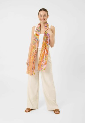 Femme souriante debout portant un débardeur blanc, un pantalon beige à jambes larges, des sandales marron et une longue écharpe colorée à motif cachemire.