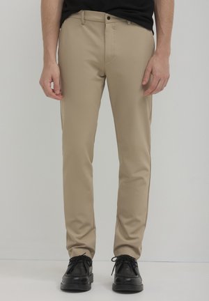 Beige Hose mit schmaler Passform, aus glattem Stoff. Verfügt über einen Knopfverschluss, Gürtelschlaufen und leicht zulaufende Beine. Getragen mit schwarzen Schuhen.