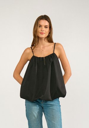 Femme aux longs cheveux portant un top noir ample à fines bretelles et un jean bleu, debout, les mains dans le dos.