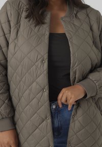 Giacca trapuntata a lunghezza ginocchio in verde oliva con bottoni automatici, polsini a costine e una finitura liscia e testurizzata. Indossata sopra una maglia nera e jeans.