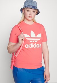 Korallkrāsas Adidas T-krekls ar baltu logo un trim svītrām. Modelis nēsā zilu ķiveres cepuri un tur rozā siksniņu somu pār ziliem spīdīgiem biksēm.