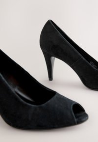 Czarne zamszowe buty na obcasie z odkrytymi palcami, smukłym obcasem i gładką teksturą. Charakteryzują się minimalistycznym wzornictwem i błyszczącym akcentem na obcasie.