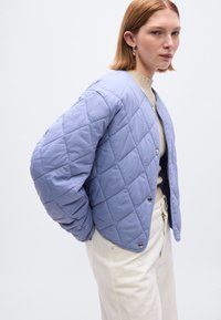 Μπουφάν σε απαλό μοβ χρώμα με καπιτονέ σχέδιο, κουμπιά με κλικ, στρογγυλεμένος λαιμός και υφή υφάσματος, συνδυασμένο με παντελόνια σε εκρού απόχρωση.