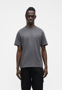 Szary bawełniany t-shirt z krótkimi rękawami i okrągłym dekoltem, na lewej piersi małe haftowane "II", noszony z czarnymi spodniami.