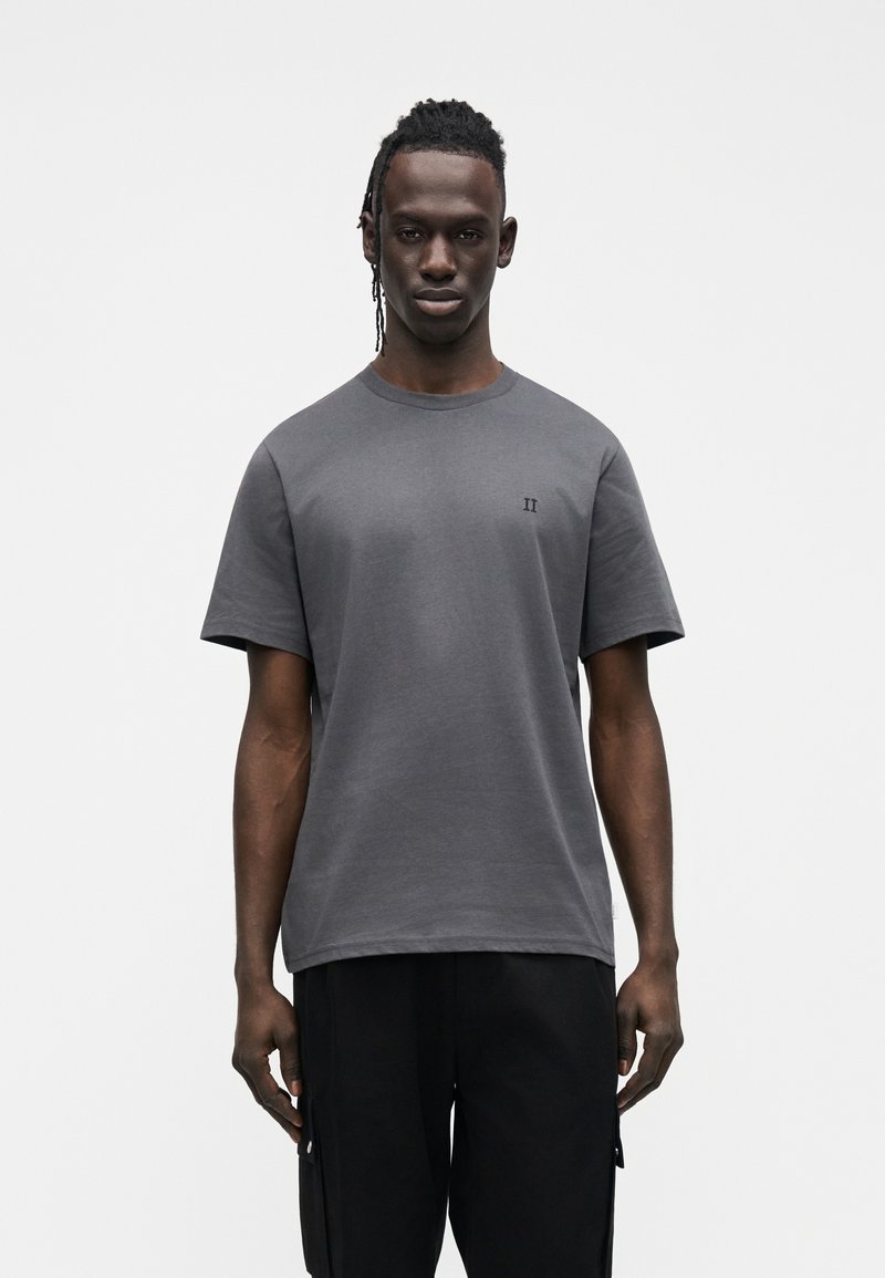 Szary bawełniany t-shirt z krótkimi rękawami i okrągłym dekoltem, na lewej piersi małe haftowane "II", noszony z czarnymi spodniami.