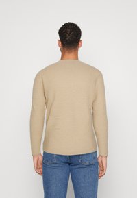 Beige tekstureret sweater med lange ærmer, rund halsudskæring, tætsiddende design og diskret mønster, parret med blå denim jeans.