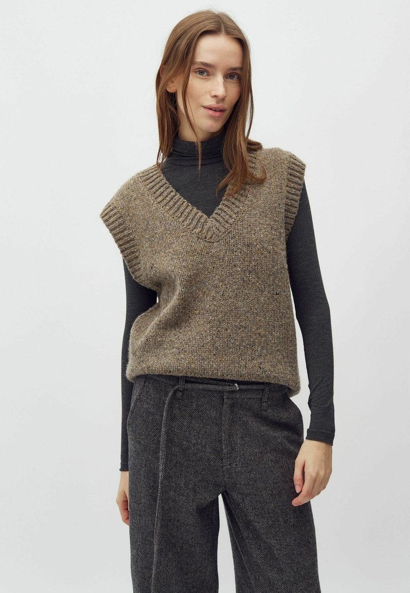 Vest tricotat cu modele maro, cu decolteu în V și margini cu nervuri, purtat peste un top negru cu mânecă lungă; asortat cu pantaloni din tweed gri.