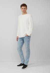 Weißes Langarmhemd, hellblaue Jeans und schwarze Sneaker. Das Model steht mit den Händen in den Taschen, die Haare sind lockig, neutraler Hintergrund.