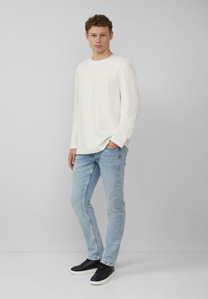 Weißes Langarmhemd, hellblaue Jeans und schwarze Sneaker. Das Model steht mit den Händen in den Taschen, die Haare sind lockig, neutraler Hintergrund.