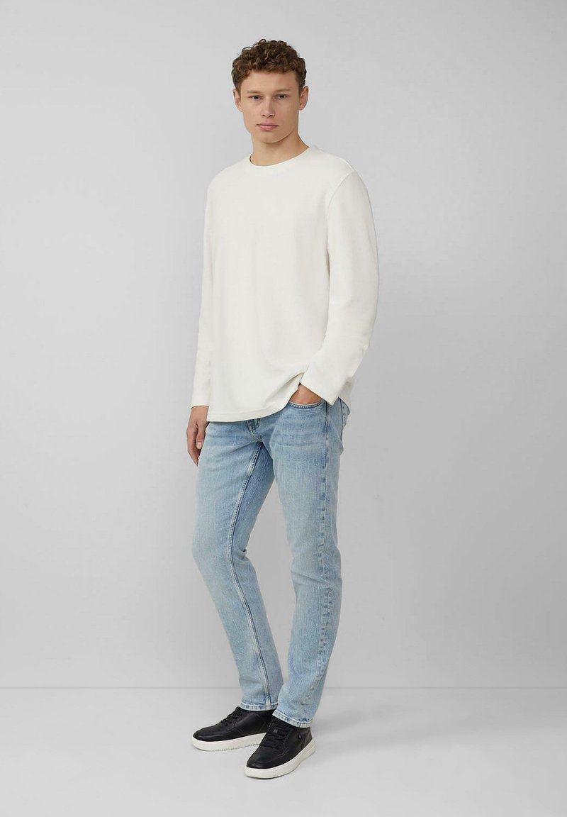 Weißes Langarmhemd, hellblaue Jeans und schwarze Sneaker. Das Model steht mit den Händen in den Taschen, die Haare sind lockig, neutraler Hintergrund.