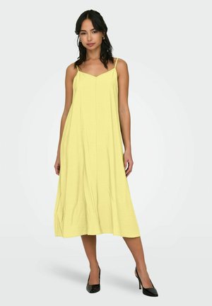 Abito midi giallo senza maniche con scollatura a V, tessuto morbido e silhouette fluida, abbinato a scarpe con tacco alto nere.
