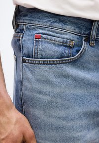 Κοντινό πλάνο τσέπης από μπλε τζιν με πορτοκαλί ραφές και μια μικρή κόκκινη ετικέτα με την επιγραφή "HUGO", με ένα χέρι να ακουμπά κοντά.