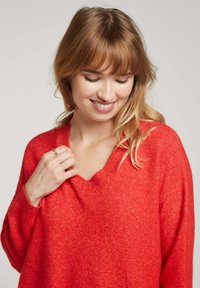 Pull en tricot doux rouge avec un décolleté en V, des épaules tombantes et des manches longues. Le matériau semble confortable avec une finition légèrement texturée.
