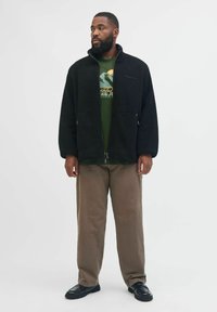 Schwarze Fleece-Zipjacke mit hohem Kragen, vorderen Taschen; getragen über einem grünen Grafik-T-Shirt, kombiniert mit beigen Hosen und schwarzen Schuhen.