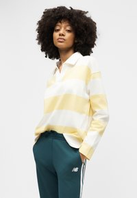 Νεαρό άτομο με κατσαρά μαλλιά, ντυμένο με μακρυμάνικη πόλο μπλούζα με κίτρινες και άσπρες ρίγες και τιρκουάζ φόρμα New Balance, με τα χέρια στις τσέπες.