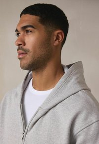 Jasnoszara bluza z zamkiem na całej długości, z dużym kapturem, o gładkiej fakturze i subtelnych detalach przeszycia, noszona na białej koszulce z okrągłym dekoltem.