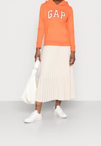 Sudadera naranja con el logo "GAP", falda plisada crema, zapatillas blancas y un bolso blanco de hombro. Tejido suave y corte relajado.