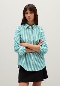 Camicia button-up a righe verticali verde petrolio e bianche, con colletto, maniche lunghe e orlo arrotondato, abbinata a una gonna nera.