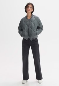 Gequilted bomberjack in gedempt grijs, met een ronde kraag en rits sluiting. Gecombineerd met zwarte broek met wijde pijpen en witte sneakers.