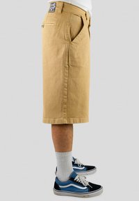Beige denimshorts med rett ben design, to frontlommer og en baklomme. Kombinert med svarte og blå joggesko og hvite sokker.