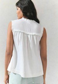 Blouse blanche sans manches avec encolure froncée et corps texturé ; s'écoule librement avec un dos froncé ; associée à un jean en denim clair.