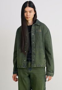 Jachetă din denim verde cu inchidere cu nasturi și două buzunare laterale, asortată cu pantaloni din denim verde. Are un finisaj texturat și o guler.