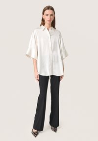 Camicia di raso bianco con collo a camicia, maniche oversize, bottoni sul davanti. Abbinata a pantaloni neri e tacchi con fantasia.