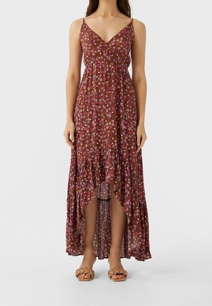 Femme portant une robe marron à fleurs sans manches avec un col en V, un ourlet asymétrique et des sandales à brides tressées sur un fond uni.