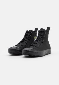 Zapatillas altas negras de lona con una puntera de goma, suela texturizada y un parche de logo redondo en verde y blanco en el lateral.