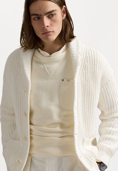 Cardigan alb tricotat cu un model texturat, guler mare de tip șal și nasturi, purtat peste un hanorac alb-cremos cu detalii la buzunar.