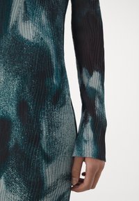Gros plan d’une main reposant près de la hanche, portant un vêtement sombre et texturé à manches longues avec des motifs abstraits turquoise et gris.