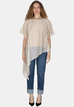 Donna con capelli ricci che indossa una maglia beige asimmetrica trasparente, jeans blu risvoltati e tacchi alti neri, in piedi davanti a uno sfondo semplice.