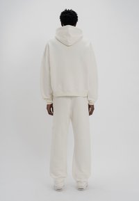 Κρεμ μα Hoodie με με�γάλο καπέλο και χαμηλωμένα ώμους, συνδυασμένο με αντίστοιχες χαλαρές φόρμες, κατασκευασμένο από απαλό υφασμάτινο υλικό.