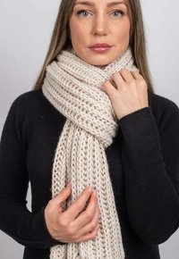 Tykt strikket tørklæde i cremefarve med et tekstureret mønster, draperet omkring halsen, holdt af hænder, mod en neutral baggrund.
