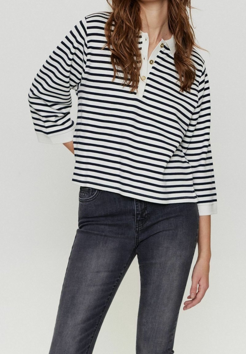 Femme portant un t-shirt à manches longues rayé blanc et marine avec des boutons dorés et un jean gris foncé, debout avec une main derrière le dos.