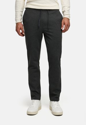 Man wearing dark gray drawstring jogger pants with white sneakers and a light long-sleeve top, standing against a plain white background.